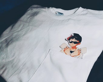 Könnte beinhalten: Weißes T-Shirt mit einer Tasche, auf der eine Grafik eines Anime-Charakters mit Sonnenbrille und Kappe abgebildet ist. Die Figur posiert mit verschränkten Armen. Das Shirt ist gefaltet und scheint aus Baumwolle zu sein.