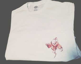 Könnte beinhalten: Weißes T-Shirt mit einer Tasche, die eine Cartoon-Geisterillustration in Rot- und Brauntönen zeigt. Der Geist hat eine Glatze und trägt eine zerfetzte Robe. Das Shirt ist auf einer grauen Oberfläche gefaltet.