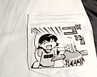 Könnte beinhalten: Hellfarbenes T-Shirt mit einer Tasche, die eine schwarz-weiße Manga-Illustration zeigt. Die Illustration enthält japanischen Text. Das T-Shirt ist gefaltet und scheint aus einem weichen Material zu sein.
