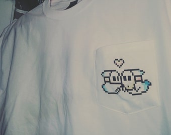 Op de afbeelding: Wit T-shirt met een zak met een gepixelde afbeelding van twee cartoonfiguren die hand in hand staan met een roze hart erboven.