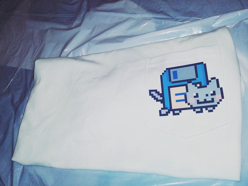May include: White t-shirt with a blue and gray pixelated cat wearing a floppy disk on its back. The cat is smiling and has a pink nose.