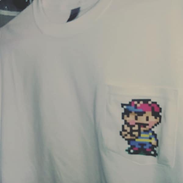 May include: White t-shirt with a pixelated image of a video game character in the pocket. The character is wearing a red hat and a blue shirt.
