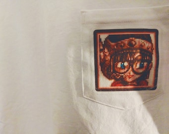 Könnte beinhalten: Weißes T-Shirt mit einer Tasche, die ein pixeliges Bild einer Figur mit Brille und Krone zeigt. Das Bild hat einen braunen Rand und ist auf dem weißen Stoff des T-Shirts platziert.