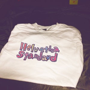May include: White long-sleeve t-shirt with a colorful graphic print that says "Helvetica Standard".