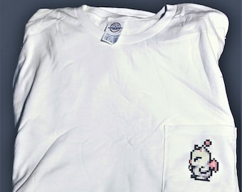 Könnte beinhalten: Weißes T-Shirt mit einem kleinen, pixeligen Bild eines gelben Wesens mit rosa Akzenten in der Tasche.