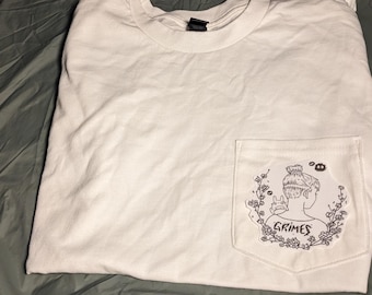 May include: White t-shirt with a pocket featuring a black and white illustration of a woman with a bun, surrounded by flowers, and the name "Grimes". The shirt is folded on a green surface.