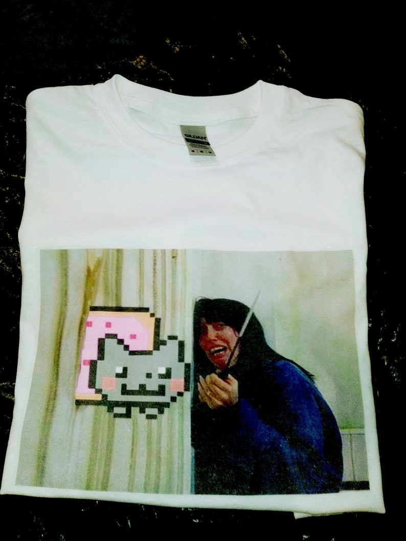 May include: White t-shirt with a printed image of a woman holding a knife and a pixelated cat with a pink and white background.