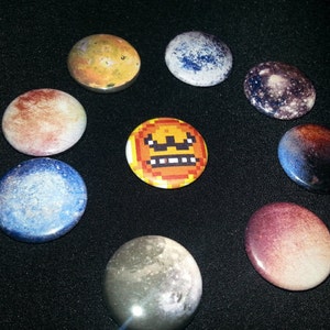 May include: A collection of round, colorful buttons with planetary and pixelated emoji designs. The buttons feature shades of blue, orange, red, and purple, with a central pixelated emoji button.