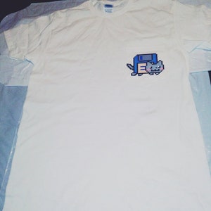 May include: White t-shirt with a pixelated image of a blue and white character holding a gray cat on the left chest.