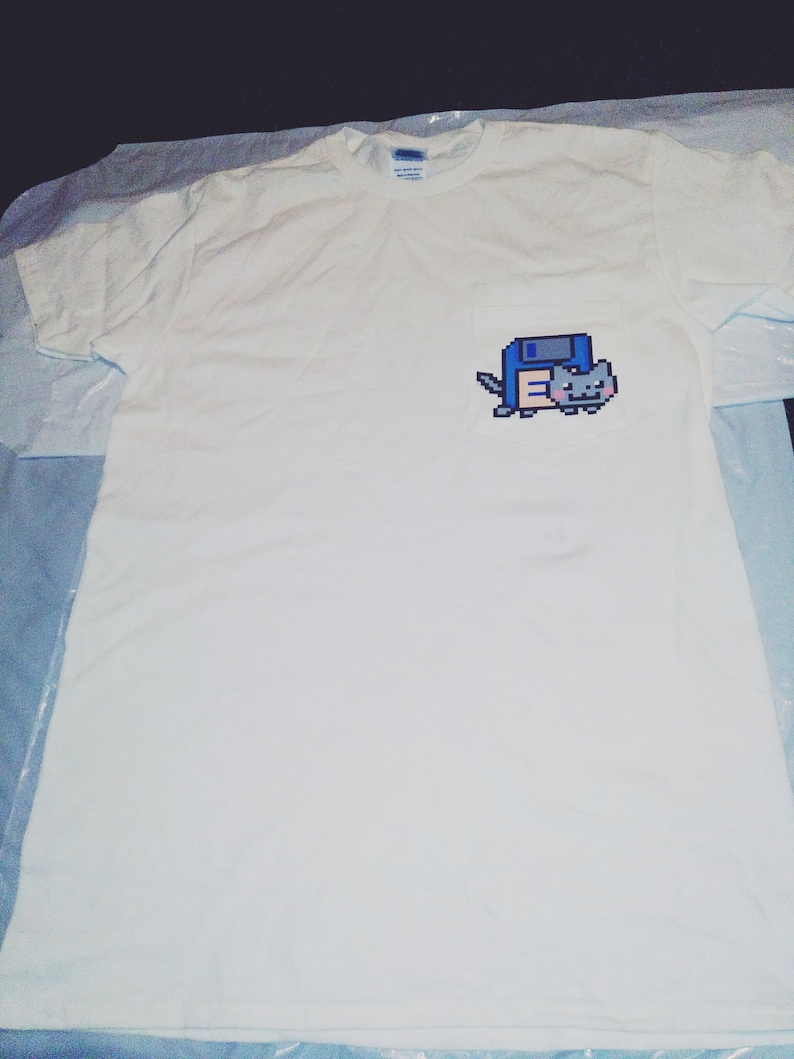 May include: White t-shirt with a pixelated image of a blue and white character holding a gray cat on the left chest.