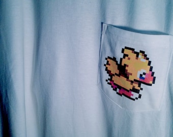 Könnte beinhalten: Hellblaues T-Shirt mit einer Tasche, die eine pixelige Cartoonfigur in Gelb, Orange, Rosa und Blau zeigt. Die Tasche befindet sich auf der linken Seite des Shirts, das aus einem weichen, hellen Stoff besteht.
