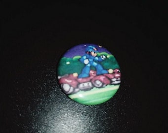 Op de afbeelding: Een ronde knop met een blauwe en paarse achtergrond waarop een cartoonfiguur op een voertuig rijdt. Het figuur is blauw en heeft een helm. Het voertuig is rood en heeft een geel licht aan de voorkant.