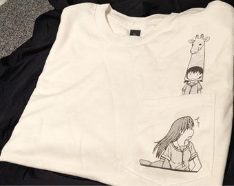 May include: White t-shirt with a pocket featuring a black and white anime illustration. The illustration includes a giraffe-like figure and a person. The shirt is folded on a dark surface.
