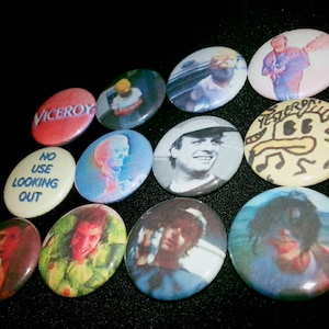 May include: A collection of 11 round pins with various designs. The pins feature images of people, cartoon characters, and text. The pins are colorful and have a variety of styles.