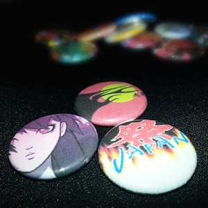 May include: Three round pinback buttons with different designs. The first button has a red background with a black silhouette of a tree and a yellow circle in the center. The second button has a purple background with a cartoon drawing of a woman with long purple hair. The third button has a white background with a red and yellow flame design and the word "JAPAN" in blue letters.