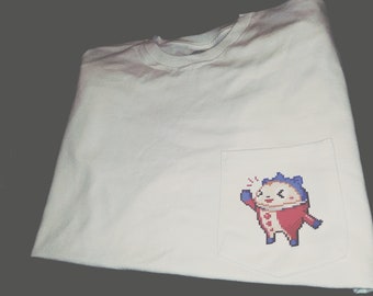 Op de afbeelding: Wit T-shirt met een zak met daarop een gepixelde cartoonfiguur in een rood-blauwe outfit. Het figuur zwaait met een verraste uitdrukking.