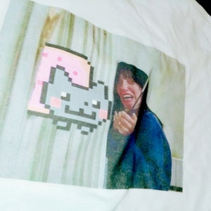 May include: White t-shirt with a printed image of a pixelated cat wearing a pink slice of bread. The image also includes a woman with long dark hair, wearing a blue shirt, looking surprised.