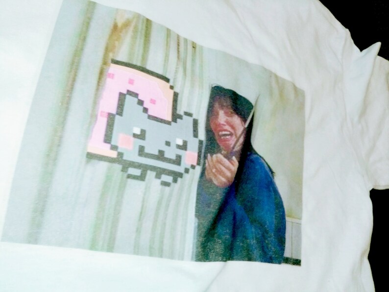 May include: White t-shirt with a printed image of a pixelated cat wearing a pink slice of bread. The image also includes a woman with long dark hair, wearing a blue shirt, looking surprised.