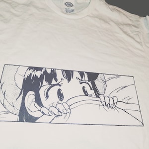 May include: A white t-shirt with a black and white graphic print of an anime character with large eyes peeking out from under a blanket.