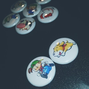 May include: A set of nine round, white button pins with various cartoon characters. The characters include a cow, a dog, a girl with a green hat, a girl with a red bow, and a boy with a green hat.