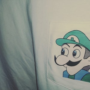 May include: A light blue shirt with a white pocket featuring a cartoon illustration of Luigi from the Super Mario Bros. series. Luigi is depicted with a green hat, blue eyes, and a large mustache. The shirt's fabric is slightly wrinkled.