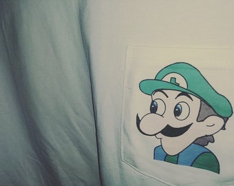 May include: A light blue shirt with a white pocket featuring a cartoon illustration of Luigi from the Super Mario Bros. series. Luigi is depicted with a green hat, blue eyes, and a large mustache. The shirt's fabric is slightly wrinkled.
