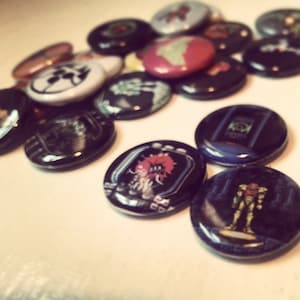 May include: A collection of colorful and black and white button pins with various designs. Some of the designs include a skull, a creature, and a video game character.
