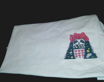 Op de afbeelding: Wit T-shirt met een zak met een kleurrijke afbeelding van een schedel met een bloemenkrans en een masker van harten en bloemen.