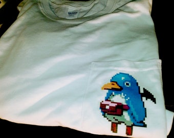 May include: White t-shirt with a pixelated penguin graphic on the pocket. The penguin is blue, with a yellow beak and holding a pink object. The shirt is folded and appears to be made of cotton.