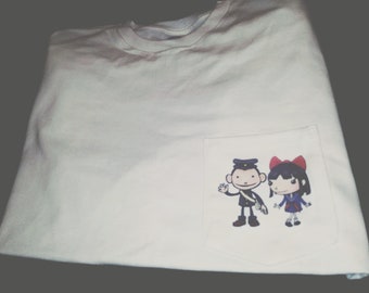 Op de afbeelding: Wit T-shirt met een zak met een cartoonillustratie van een jongen en een meisje. De jongen draagt een blauw uniform en een hoed, en het meisje draagt een blauwe jurk en een rode strik.