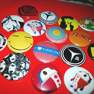 May include: A collection of 14 round buttons with various designs. The buttons are colorful and feature images of cartoon characters, geometric shapes, and text. The buttons are arranged in a circle on a red background.
