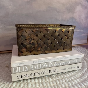 May include: A rectangular brass basket with a woven lattice design and a textured rim, resting on a stack of three books titled The Story of American Painting, Billy Baldwin decorates, and Memories of Home by Heidi Caillier.