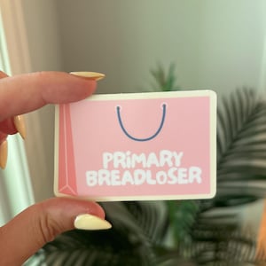 May include: A pink rectangular sticker with a shopping bag design. The sticker has a white border and the words "PRIMARY BREADLOSER" in white block letters. A blue handle is at the top.