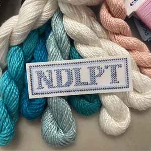 Puede incluir: Una colecci&oacute;n de hilos de bordar en varios colores, incluyendo blanco, turquesa y rosa. Tambi&eacute;n se ve una etiqueta con las letras "NDLPT". Los hilos est&aacute;n cuidadosamente dispuestos, sugiriendo un suministro de manualidades.