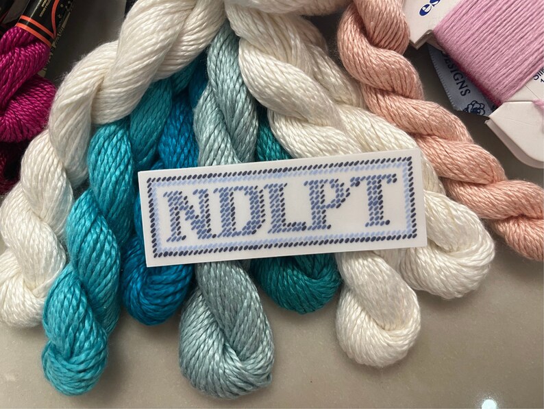 Puede incluir: Una colecci&oacute;n de hilos de bordar en varios colores, incluyendo blanco, turquesa y rosa. Tambi&eacute;n se ve una etiqueta con las letras "NDLPT". Los hilos est&aacute;n cuidadosamente dispuestos, sugiriendo un suministro de manualidades.