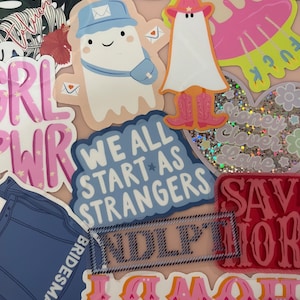 Puede incluir: Una colecci&oacute;n de pegatinas coloridas sobre una superficie rosa claro. Las pegatinas presentan varios dise&ntilde;os, incluyendo un fantasma con sombrero y botas de vaquero, un fantasma con gorra de cartero, y el texto "We All Start As Strangers."