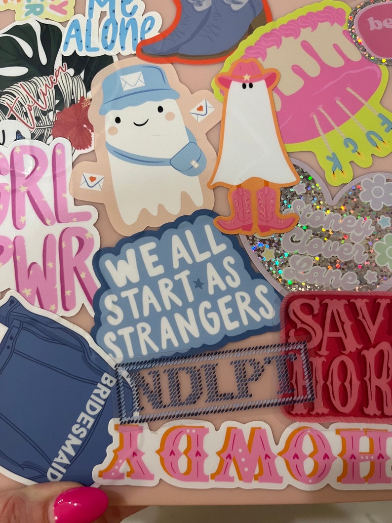 Puede incluir: Una colecci&oacute;n de pegatinas coloridas sobre una superficie rosa claro. Las pegatinas presentan varios dise&ntilde;os, incluyendo un fantasma con sombrero y botas de vaquero, un fantasma con gorra de cartero, y el texto "We All Start As Strangers."