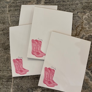 May include: Four white notepads with pink cowboy boots and the word "howdy" written on each notepad.