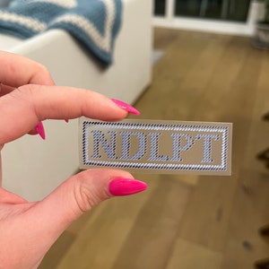Puede incluir: Una pegatina rectangular con las letras "NDLPT" en azul claro, con una fuente estilizada. Las letras est&aacute;n sobre un fondo transparente, enmarcadas por un borde blanco y negro similar a una cuerda.