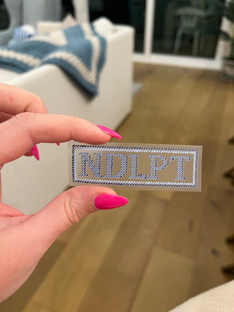 Puede incluir: Una pegatina rectangular con las letras "NDLPT" en azul claro, con una fuente estilizada. Las letras est&aacute;n sobre un fondo transparente, enmarcadas por un borde blanco y negro similar a una cuerda.