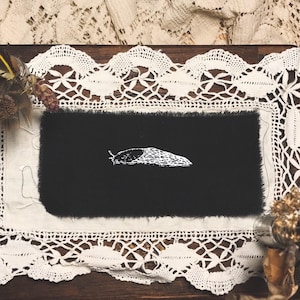 May include: A black fabric square with a white embroidered slug on a white lace doily. The doily is on a wooden surface. There are feathers and bones around the doily.