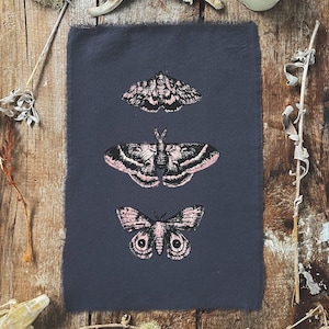 Puede incluir: Una tela gris oscura con tres ilustraciones de polillas negras y blancas impresas en ella. Las polillas tienen acentos rosados. La tela se muestra sobre una superficie de madera con ramas secas y un hueso.