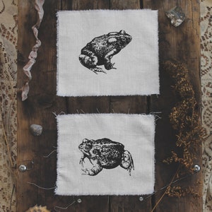 May include: Two white fabric squares with black and white illustrations of frogs. The squares are on a wooden surface with a brown lace fabric on the left and a brown fern on the right.