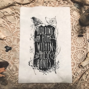 Könnte beinhalten: Schwarz-weiß-Illustration eines Grabsteins mit dem Text "Building a home within these bones" auf einem weißen Stoffhintergrund.