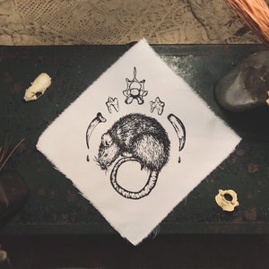 Peut inclure: Une illustration en noir et blanc d'un rat enroulé sur un fond de tissu blanc. Le rat est entouré d'os et de dents, créant une esthétique mystique et macabre.