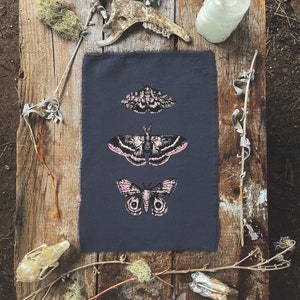 May include: A dark gray fabric with three black and white moth illustrations printed on it. The moths are in a vertical line, with the largest moth at the bottom. The fabric is surrounded by natural elements, including branches, moss, and bones.