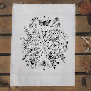 Könnte beinhalten: Weißes Leinen-Geschirrtuch mit einer schwarz-weißen botanischen Illustration. Das Design zeigt einen zentralen Schädel, umgeben von Blumen, Insekten und anderen natürlichen Elementen. Das Tuch wird auf einer Holzoberfläche präsentiert.