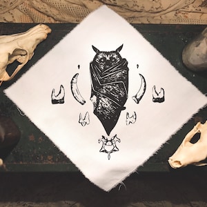 May include: A white fabric square featuring a black and white illustration of a bat with its wings outstretched. The bat is surrounded by a symmetrical arrangement of stylised teeth and fangs.
