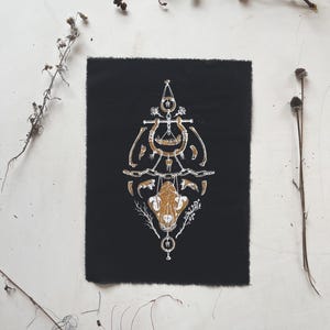 Pode incluir: Um remendo de tecido preto com um design branco e dourado de um crânio de animal com ossos e outros elementos. O design é simétrico e tem uma sensação mística ou ocultista.