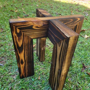 May include: A small, handcrafted wooden plant stand with a unique geometric design. The stand is made of dark-stained wood, showcasing the natural grain and knots. The legs are angled, providing stability. The stand is suitable for displaying plants or decorative items.